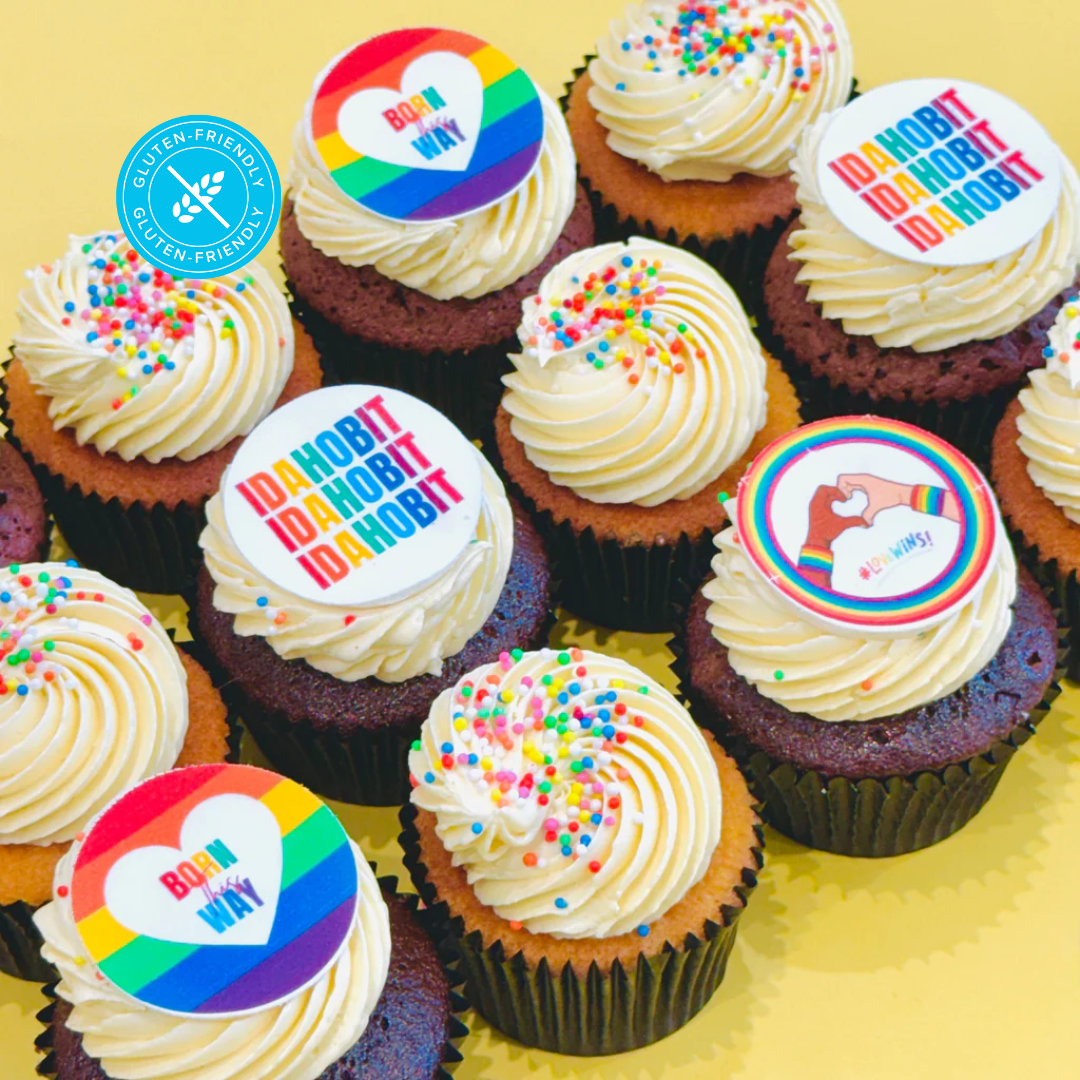 Gluten-friendly cupcakes with vanilla frosting, colourful IDAHOBIT toppers, and sprinkles, arranged on a yellow background