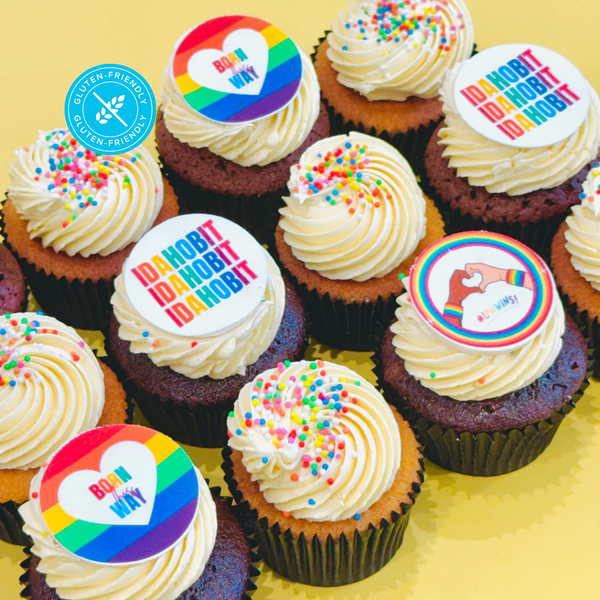Gluten-friendly cupcakes with vanilla frosting, colourful IDAHOBIT toppers, and sprinkles, arranged on a yellow background