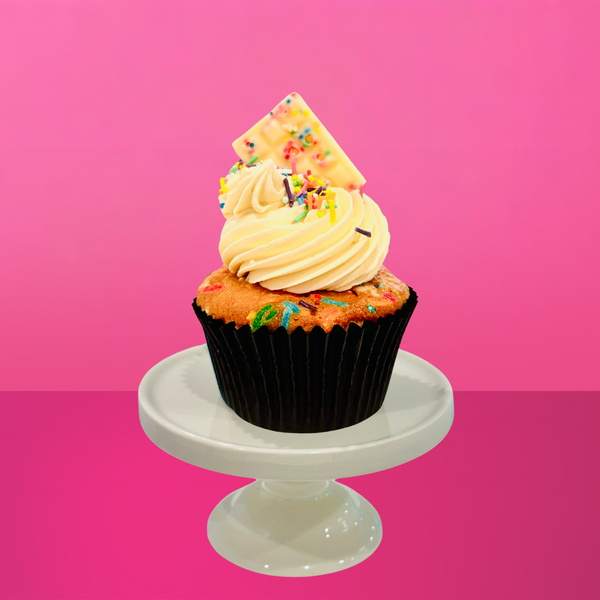 Rainbow sprinkle cupcake with vanilla buttercream and a gold chocolate bar on a pink backdrop.