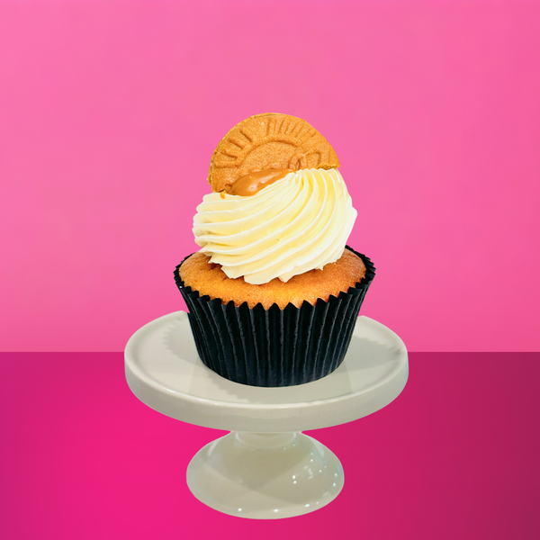 Vanilla cupcake with piped vanilla buttercream, cookie butter drizzle and a Biscoff cookie on top, displayed on a white stand against a pink background.