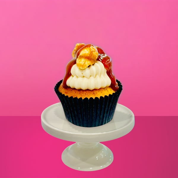 Caramel cupcake topped with piped vanilla buttercream, a drizzle of salted caramel sauce, and caramel popcorn pieces, sitting on a white mini cake stand against a hot pink background.