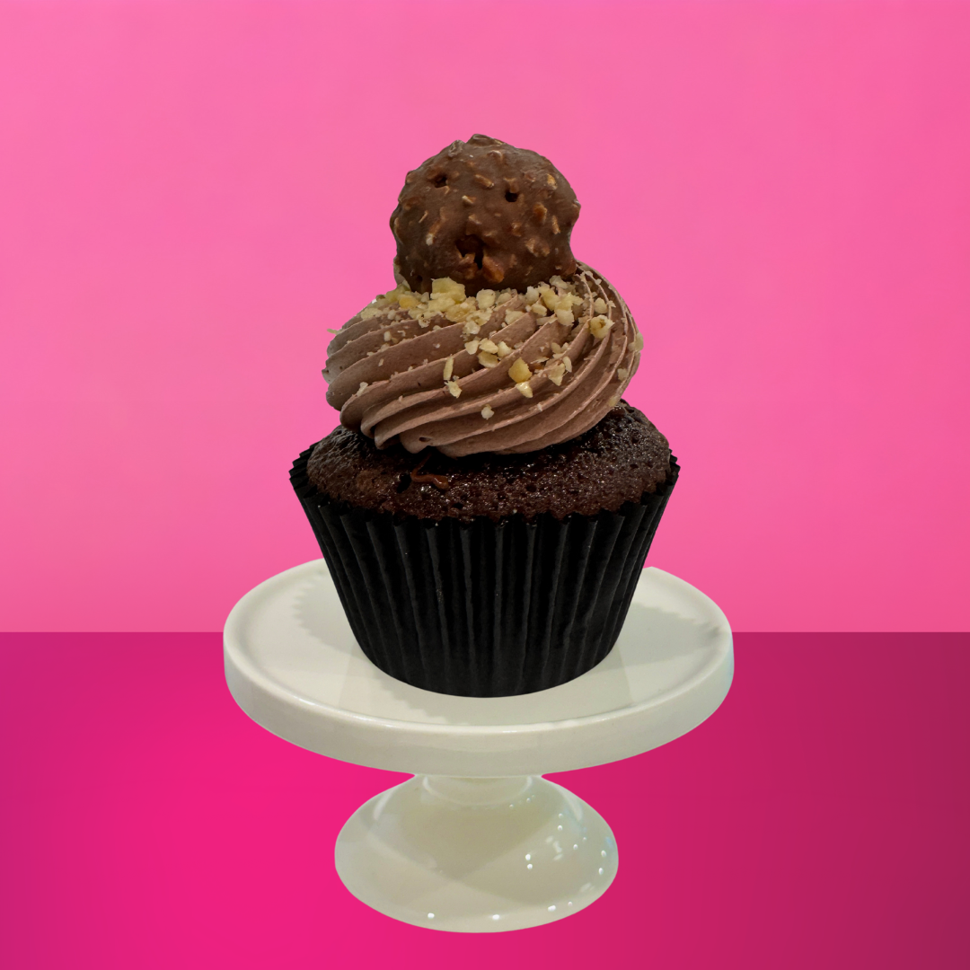 Chocolate fudge cupcake topped with swirls of milk chocolate buttercream, crushed hazelnuts, and a Ferrero-style chocolate truffle on top, displayed on a white cake stand with a vibrant pink background.