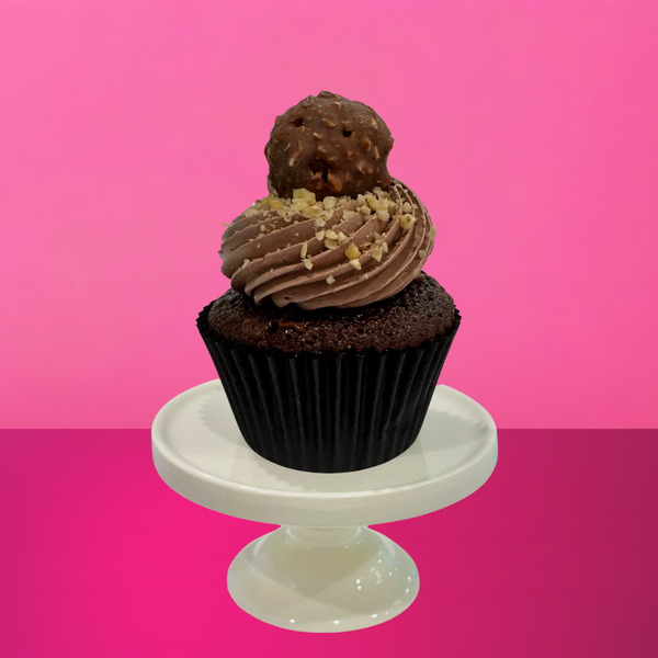 Chocolate fudge cupcake topped with swirls of milk chocolate buttercream, crushed hazelnuts, and a Ferrero-style chocolate truffle on top, displayed on a white cake stand with a vibrant pink background.