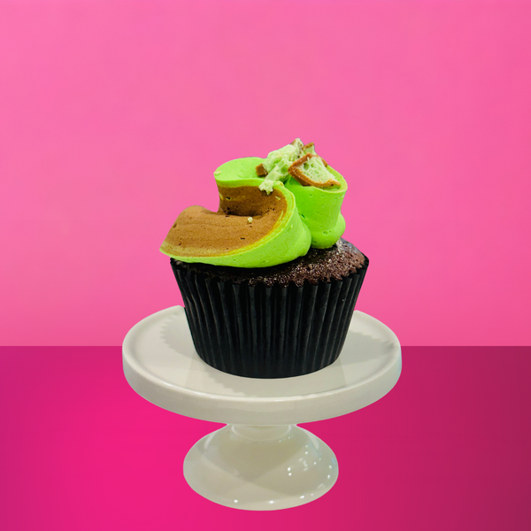 Chocolate cupcake topped with a swirl of green and brown mint-chocolate buttercream and a piece of mint chocolate bar, displayed on a small white cake stand against a vibrant pink background.