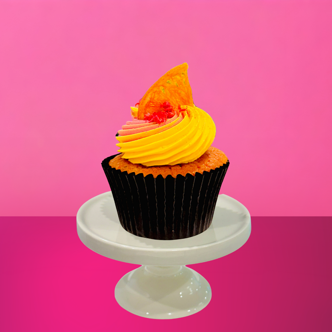 Pink and yellow frosted cupcake topped with a dried citrus slice and a sprinkle of crushed candy, displayed on a white mini cake stand against a bold pink background.
