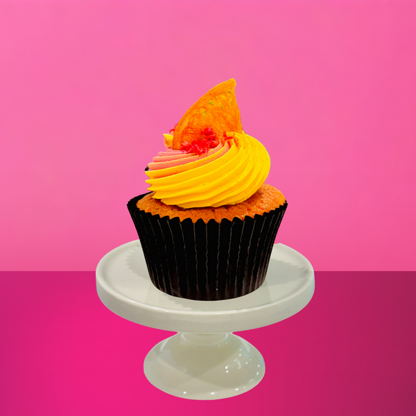 Pink and yellow frosted cupcake topped with a dried citrus slice and a sprinkle of crushed candy, displayed on a white mini cake stand against a bold pink background.
