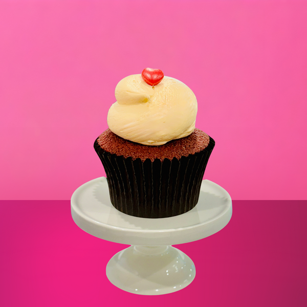 Red velvet cupcake topped with a swirl of cream cheese frosting and finished with a small red candy heart, set against a pink background.
