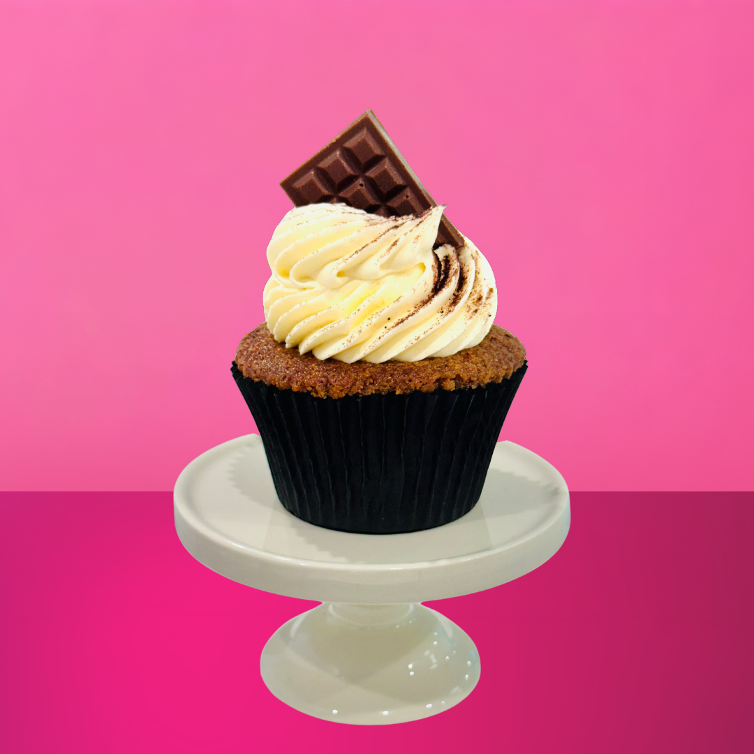 Espresso-soaked vanilla cupcake topped with mascarpone buttercream, cocoa dusting, and a mini chocolate bar on a white stand against a pink background.