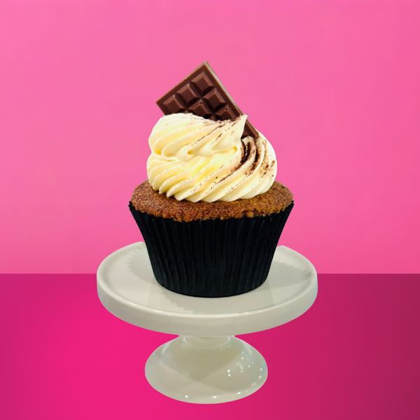 Espresso-soaked vanilla cupcake topped with mascarpone buttercream, cocoa dusting, and a mini chocolate bar on a white stand against a pink background.