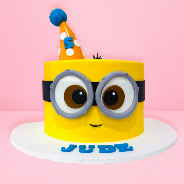 A yellow Minions-themed birthday cake with large gray and brown eyes, a black band, and a blue party hat on top, 
